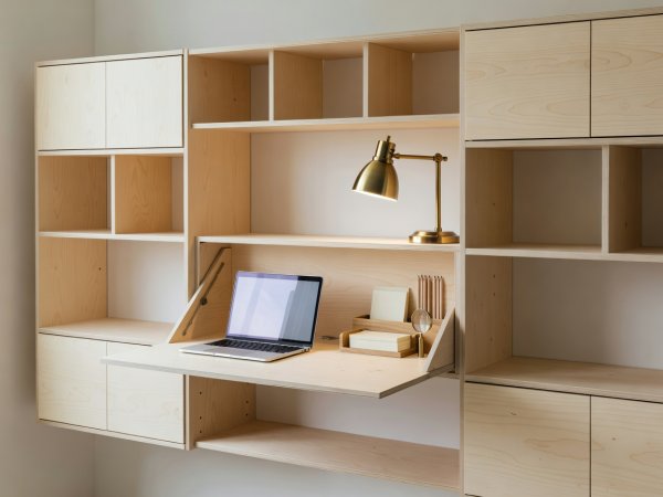 Wall-mounted birch modular shelving unit with a drop-front desk cabinet open to show a laptop, brass lamp and wooden stationery tray